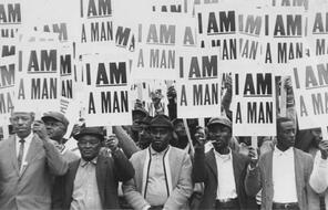Photo of Martin Luther King, Jr. marching arm in arm with a crowd of men participating in the  1968 Memphis Sanitation Worker's Strike.