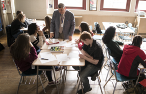 An educator stands at the head of a table filled with students in conversation.