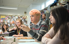 A student with pink hair speaks in a classroom with their hand raised.