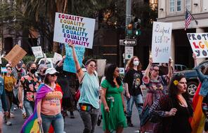 A group of people walking down a street holding signs that read "Equality" and "Defend LGBTQ+ Youth".