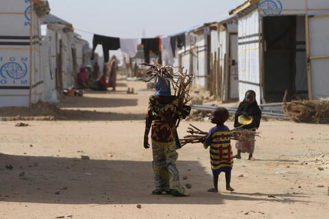 A Nigerian woman and young boy walk, holding sticks, among makeshift homes in a refugee camp.