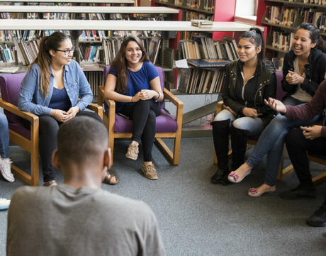 Nine students sitting in a circle in a school library, laughing and smiling
