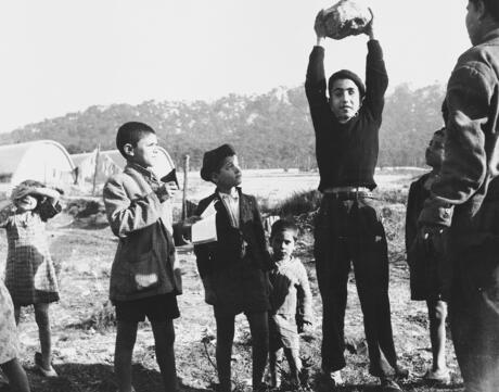 A group of boys gather in the Los Arenas camp. One boy stands in the middle holding a rock over his head while others look at him.