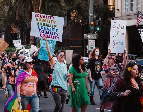 A group of people walking down a street holding signs that read "Equality" and "Defend LGBTQ+ Youth".