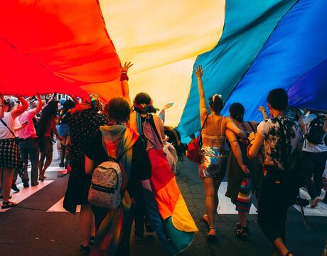 Photograph of people walking under rainbow flag during Pride parade.